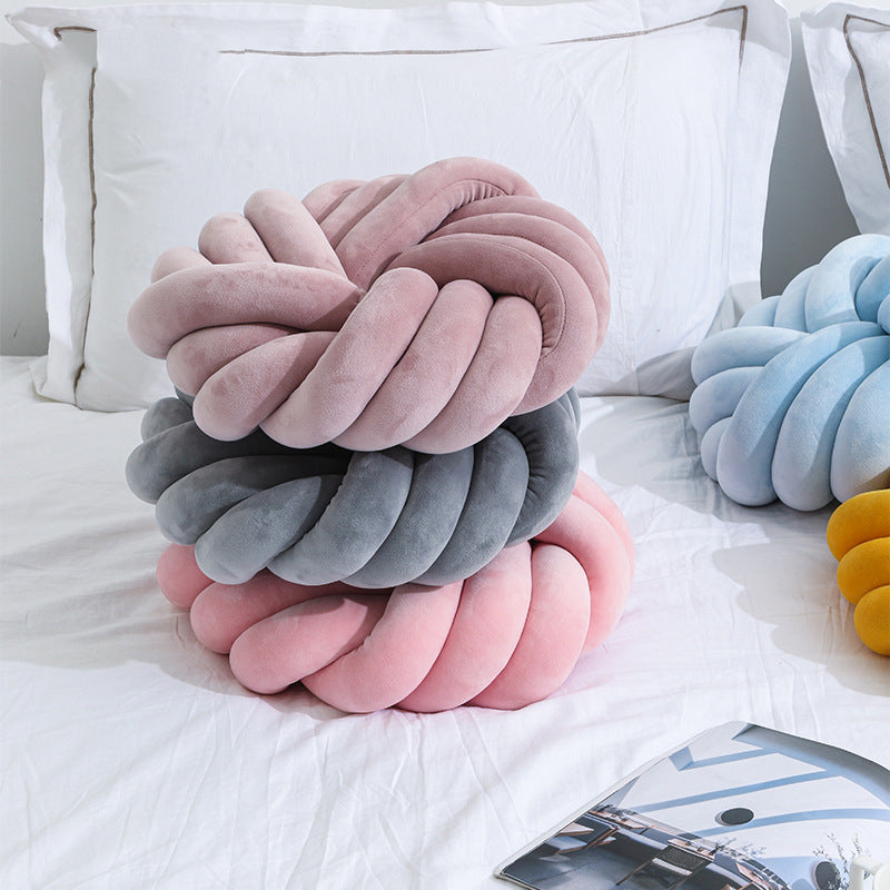 Stack of knotted cushions in pink, gray, and yellow on a bed.