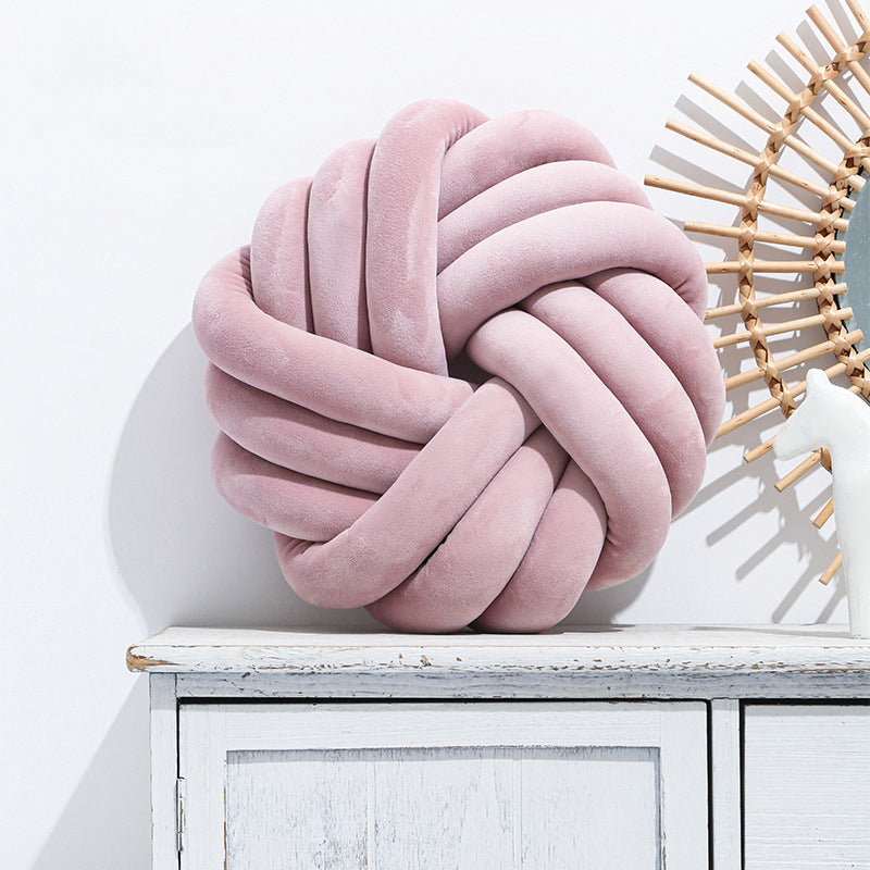 Pink knotted pillow on a white surface with a decorative item in the background
