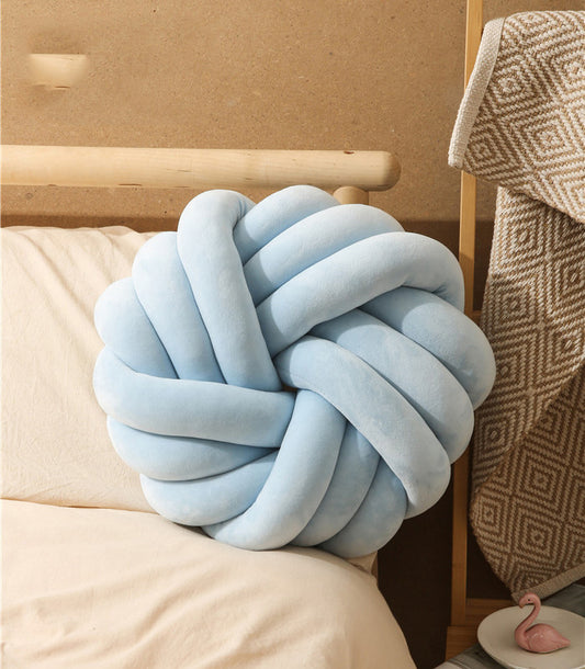 Light blue knot pillow on a bed with beige bedding and a patterned blanket.